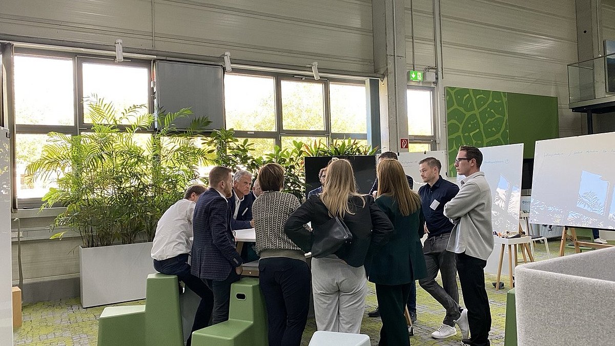 Worshopteilnehmende stehen und sitzen in einem Kreis um einen Tisch zusammen und besprechen ein Workshopthema.