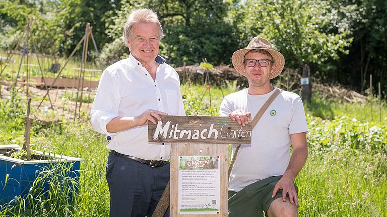 Minister Franz Untersteller posiert für ein Foto in einem Garten mit einem Mann, auf einem Schild steht "Mitmach Garten"