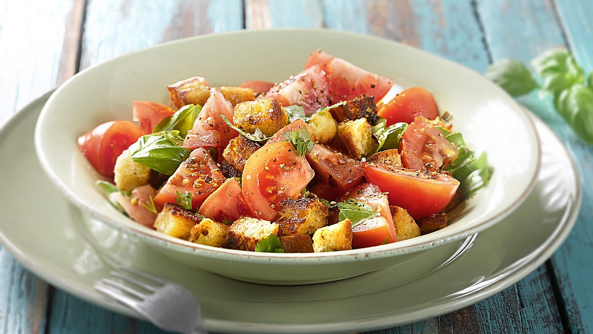 Auf einem blaugestrichenen Holztisch steht ein Teller mit Brotsalat aus Brotresten, Tomaten und Kräutern.