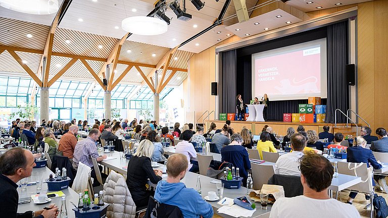 Das Bild zeigt eine Veranstaltungshalle, in der einige Personen an Tischen sitzen und den Blick auf die Bühne richten, die rechts hinten im Bild zu sehen ist. Dort stehen drei Frauen, darunter Kultusministerin Theresa Schopper und Umweltministerin Thekla Walker.