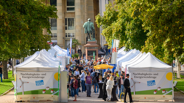 Die Aufnahme zeigt die Allee beim Schlossplatz in Stuttgart, in der Pavillons aufgebaut sind und Menschen herumlaufen. Am Anfang der Pavillons weisen zwei Schilder auf die Nachhaltigkeits- und Energiewendetage 2023 hin.