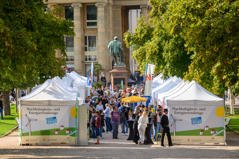 Die Aufnahme zeigt die Allee beim Schlossplatz in Stuttgart, in der Pavillons aufgebaut sind und Menschen herumlaufen. Am Anfang der Pavillons weisen zwei Schilder auf die Nachhaltigkeits- und Energiewendetage 2023 hin.