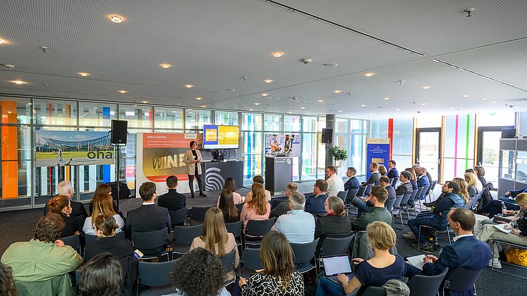 Mehrere Personen sitzen in einem Raum und hören einer Rednerin zu, die etwas vorträgt. Dahinter ist ein Plakat der WIN-Charta zu sehen.