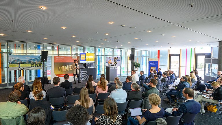 Mehrere Personen sitzen in einem Raum und hören einer Rednerin zu, die etwas vorträgt. Dahinter ist ein Plakat der WIN-Charta zu sehen.
