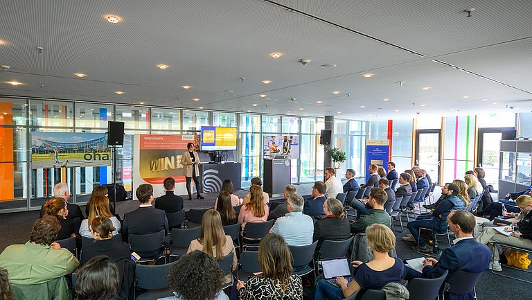 Mehrere Personen sitzen in einem Raum und hören einer Rednerin zu, die etwas vorträgt. Dahinter ist ein Plakat der WIN-Charta zu sehen.