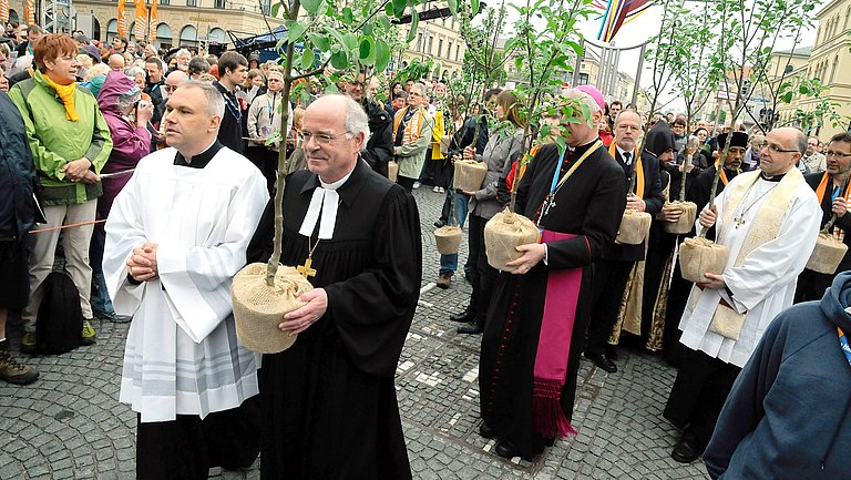 Ökomenischer Kirchentag Verschieden Kirchenvertreter schreiten durch eine Straße gesäumt von vielen Gläubigen. Einige Kirchenvertreter tragen Baumsetzlinge.