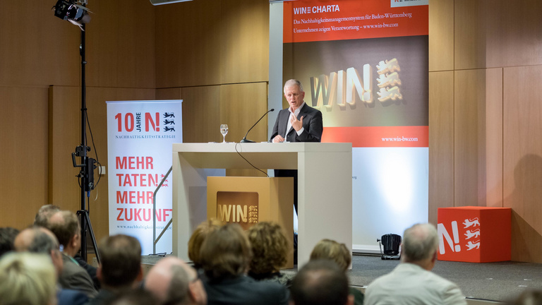 Ein Redner hält bei einer Veranstaltung der Wirtschaftsinitiative Nachhaltigkeit am Redepult eine Vortrag vor dem Publikum.