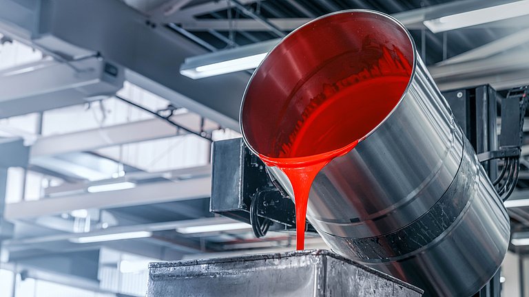 In einer hellen Produktionshalle fließt aus einem großen Metallfass rote Farbe in den Container einer Abfüllanlage.