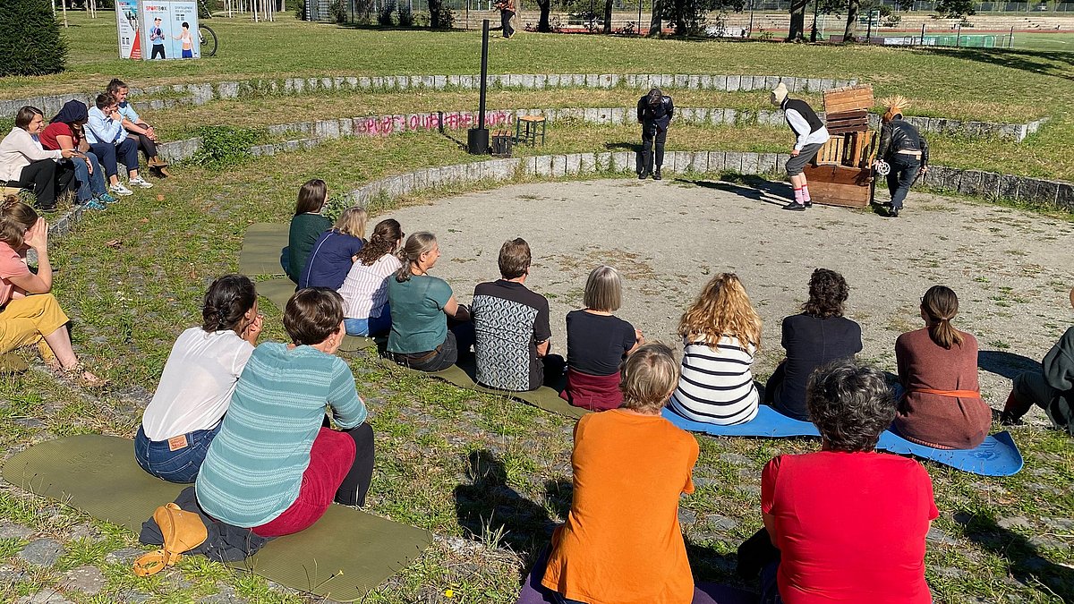Teilnehmende einer Veranstaltung sitzen in einem Freilicht-Atrium und schauen einer Theateraufführung zu.