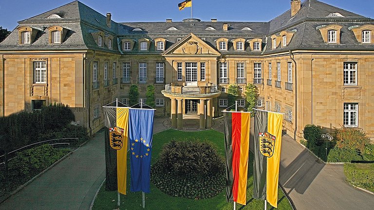 Die Villa Reitzenstein ist ein antikes, prachtvolles Gebäude mit einer Steinfassade und grauem Dach. Vor ihr sind vier verschiedene Fahnen gehisst. Unter anderem die europäische, die deutsche und die baden-württembergische Flagge. 