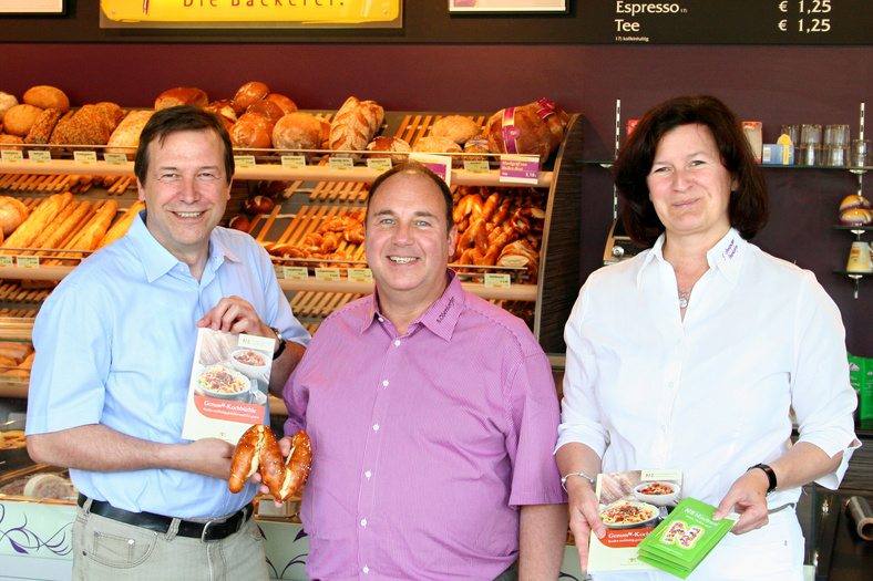 drei Mitarbeitende der Bäckerei Armbruster zeigen in die Kamera ihre gebackenen N und das Genuss-Kochbuch