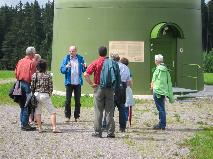 Eine Gruppe von sieben Personen vor dem grünen Sockel einer Windkraftanlage.