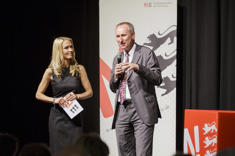 Nachhaltigkeitstage 2014 Ein Mann im Anzug steht auf der Bühne. Er redet zum Publikum, spricht ins Mikrofon. Die Moderatorin steht neben ihm und schaut ihn an.