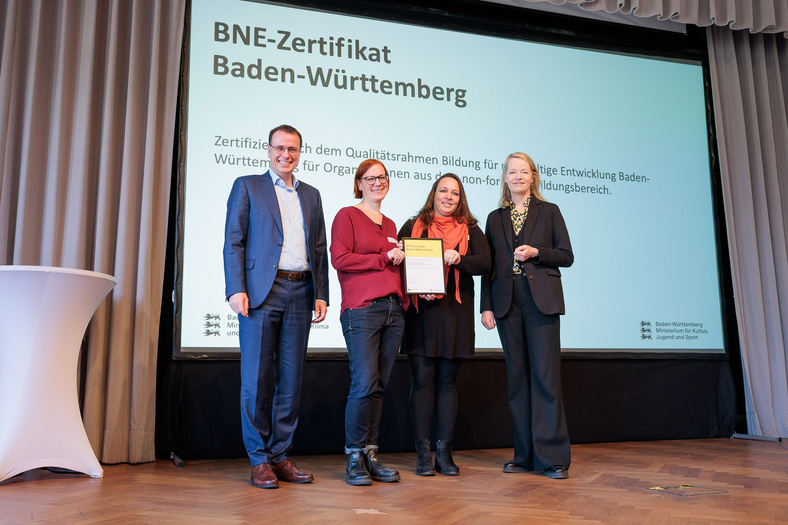 Umweltministerin Thekla Walker MdL und Kultusstaatssekretär Volker Schebesta MdL stehen zusammen mit zwei Frauen auf einer Bühne. Sie lächeln alle in die Kamera, die zwei Frauen halten ein eingerahmtes BNE-Zertifikat.