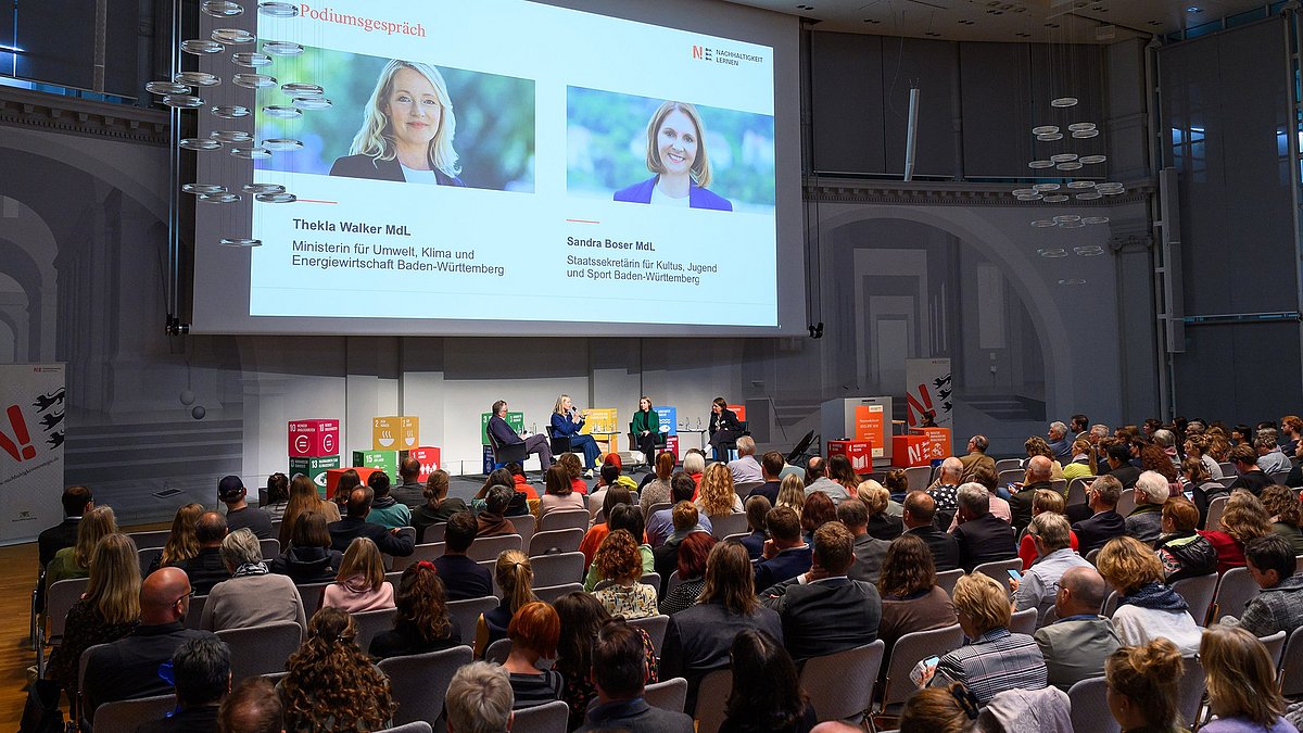 Auf der Bühne findet eine Podiumsdiskussion statt zwischen der Umweltministerin Thekla Walker und der Staatssekretärin für Kultur, Jugend und Sport in Baden-Württemberg Sandra Boser. Über Ihnen ist eine große Leinwand auf der eine Präsentation zu sehen ist. Im Vordergrund sind die mit den Zuhörenden gefüllten Stuhlreihen.