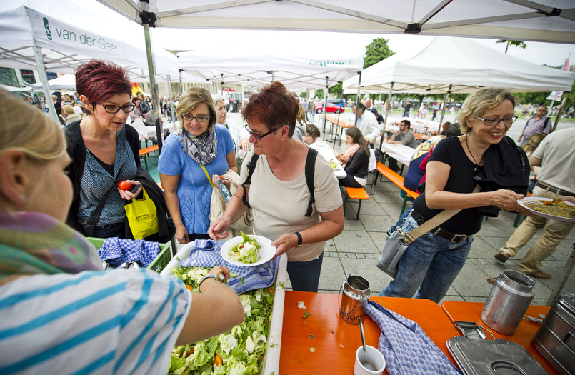 Essensausgabe auf dem Schlossplatz in Stuttgart. Eine Frau hat einen Teller mit Salat in der Hand. Bierbänke und Biertische.