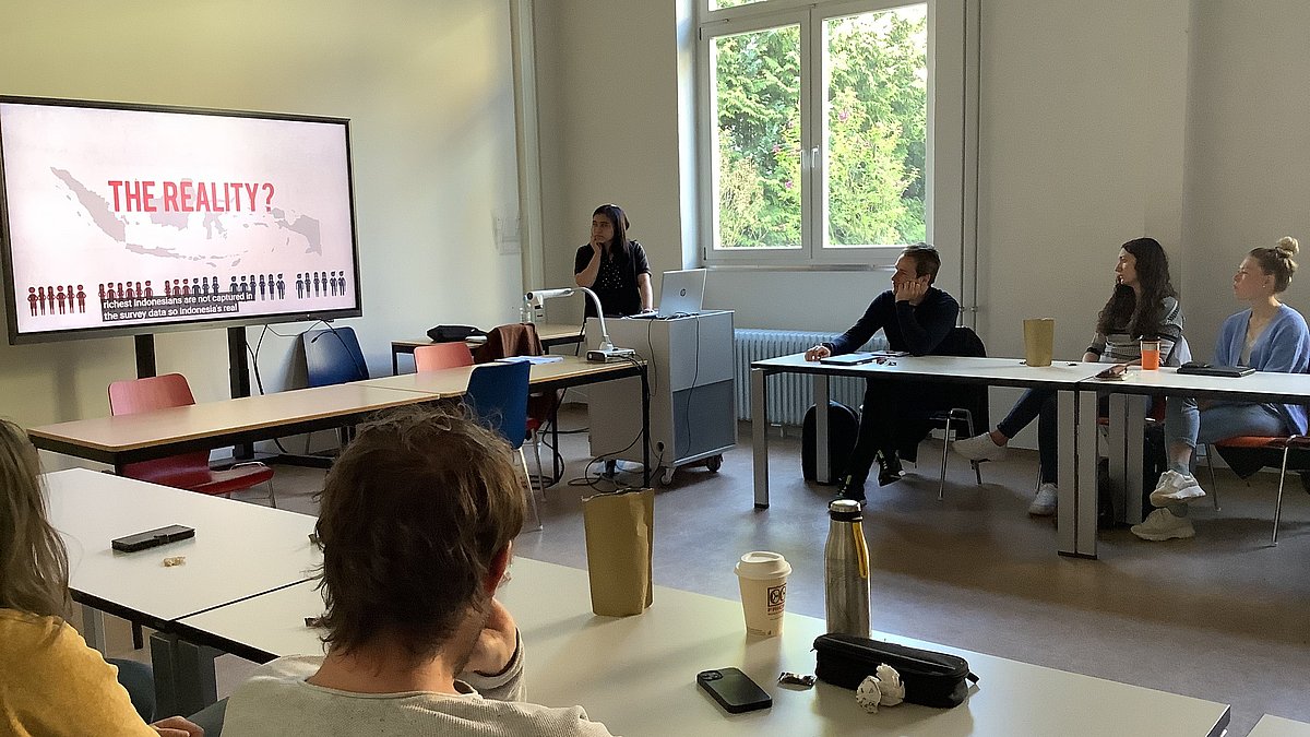 Mehrere Personen sitzen in einem Schulraum an U-förmig aufgestellten Tischen und blicken auf einen Präsentationsbildschirm, neben dem eine Frau steht und etwas vorträgt.