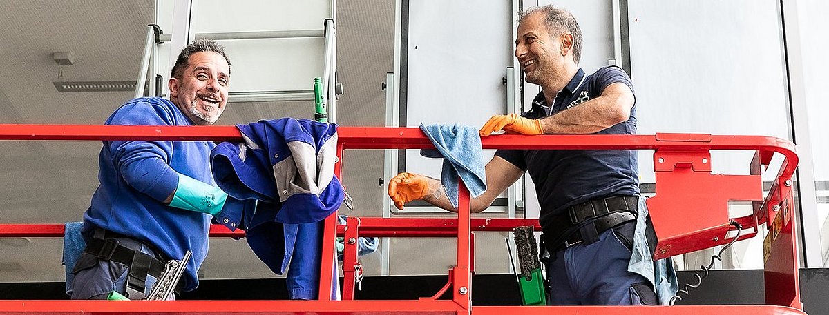 Zwei Männer stehen lachend auf einer roten Hebebühne und haben Putzutensilien dabei.