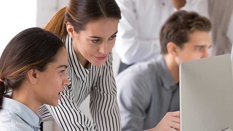 Eine Schülerin und ein Mitarbeiterin aus einem Unternehmen besprechen sich am Computer. Im Hintergrund arbeiten ein Schüler und ein Mitarbeiter zusammen.
