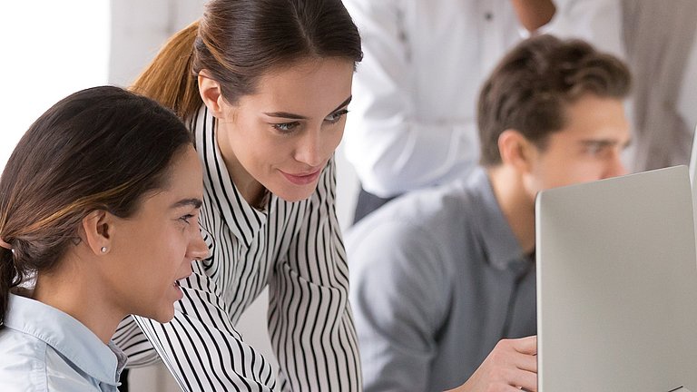 Eine Schülerin und ein Mitarbeiterin aus einem Unternehmen besprechen sich am Computer. Im Hintergrund arbeiten ein Schüler und ein Mitarbeiter zusammen.