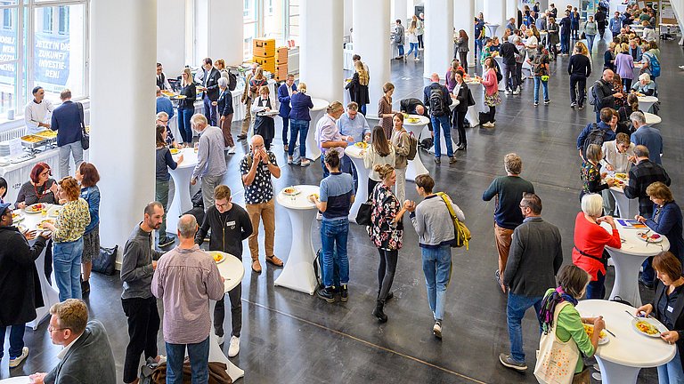 Ein Raum mit vielen runden Stehtischen ist gefüllt von Menschen, die sich gerade am Catering bedienen und ihr Mittagessen genießen.
