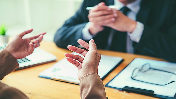 Dargestellt sind zwei Personen, von denen man jeweils nur die Hände sieht. Eine Person scheint etwas zu sagen, die andere Person sitzt gegenüber und hört ihr zu.