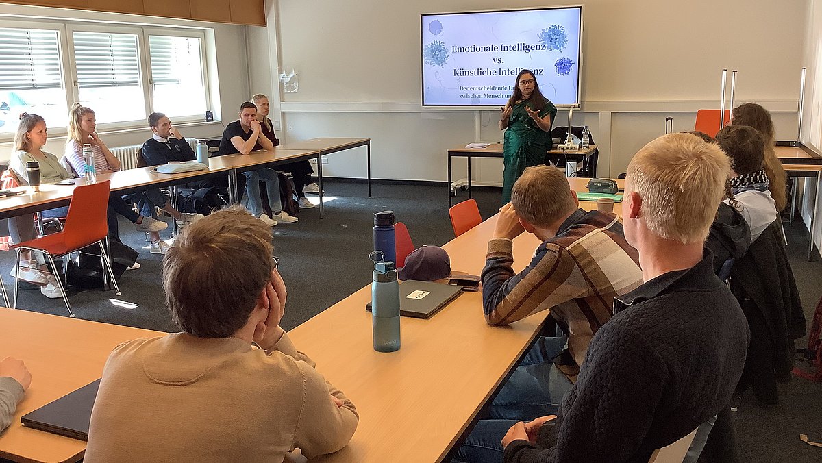 Mehrere Personen sitzen in einem Klassenraum an U-förmig aufgestellten Tischen und blicken zu einem Präsentationsbildschirm, vor dem eine Person steht und einen Vortrag hält.