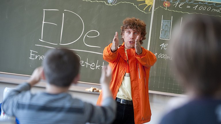 Ein Mann mit orangener Jacke erklärt Schülern in einem Klassenzimmer Dinge übers Energiesparen.