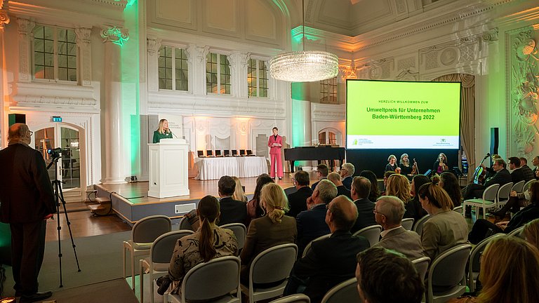 Umweltministerin Thekla Walker steht am Rednerpult auf der Bühne, auf der eine weitere Frau sowie eine Leinwand mit der Aufschrift "Umweltpreis für Unternehmen Baden-Württemberg 2022" stehen. Vor der Bühne sitzen mehrere Menschen mit Blick zur Bühne.
