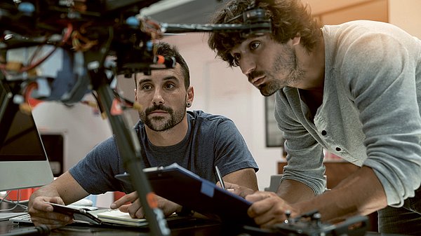 Dargestellt sind zwei Männer, die aufmerksam ein von ihnen entwickeltes Gerät betrachten.