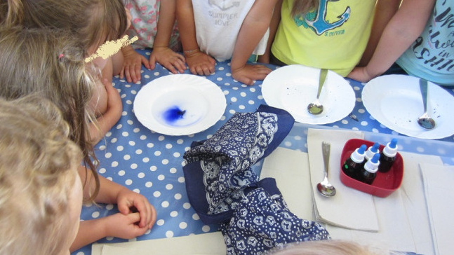 Mehrere Kinder stehen um einen Tisch, auf dem drei Teller stehen. Auf einem davon befindet sich ein blauer Eiswürfel.