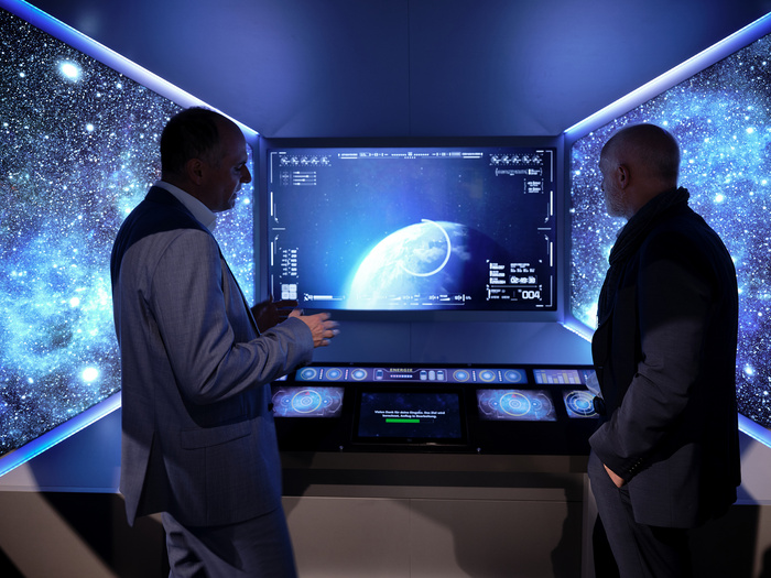 Zwei Männer stehen in einem dunklen Raum vor einem Bildschirm, der die Erde als Planeten im Weltraum zeigt.