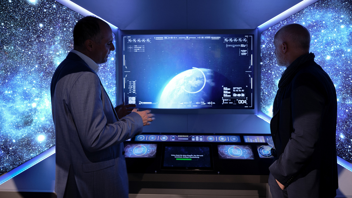 Zwei Männer stehen in einem dunklen Raum vor einem Bildschirm, der die Erde als Planeten im Weltraum zeigt.
