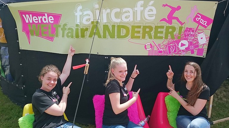 Drei junge Frauen sitzen auf grellbunten Plastikstühlen vor einem Plakat für faircafé der „WELTfairÄNDERER“.