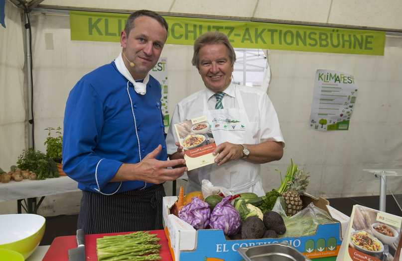 Franz Unterstelle hält das Genuss-Kochbuch der Nachhaltigkeitsstrategie in die Kamera und steht mit einem Koch vor einem Haufen Gemüse