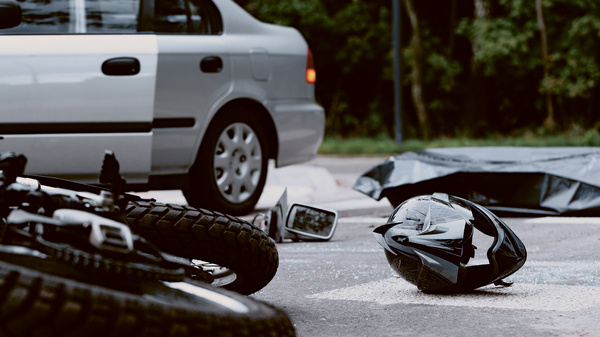 Dargestellt ist eine Unfallszene. Ein Motorrad und ein Helm liegen auf dem Boden, daneben steht ein Pkw.