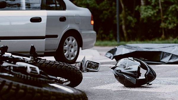 Dargestellt ist eine Unfallszene. Ein Motorrad und ein Helm liegen auf dem Boden, daneben steht ein Pkw.