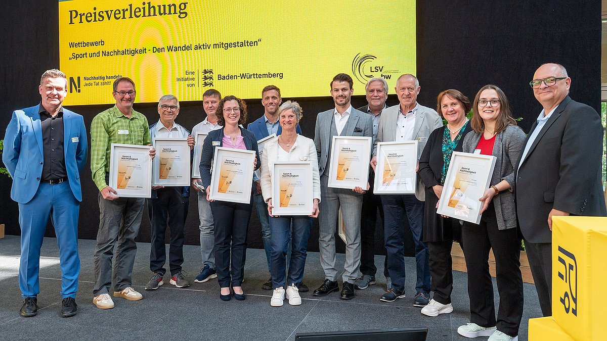 Farbige Fotografie: Gruppenbild auf der Bühne mit allen Preisträgerinnen und Preisträgern. Alle halten ihre Urkunden 'Sport & Nachhaltigkeit' in der Hand.