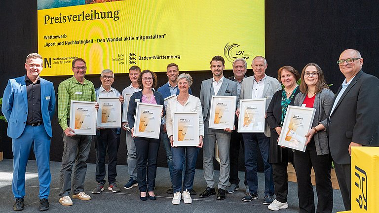 Farbige Fotografie: Gruppenbild auf der Bühne mit allen Preisträgerinnen und Preisträgern. Alle halten ihre Urkunden 'Sport & Nachhaltigkeit' in der Hand.