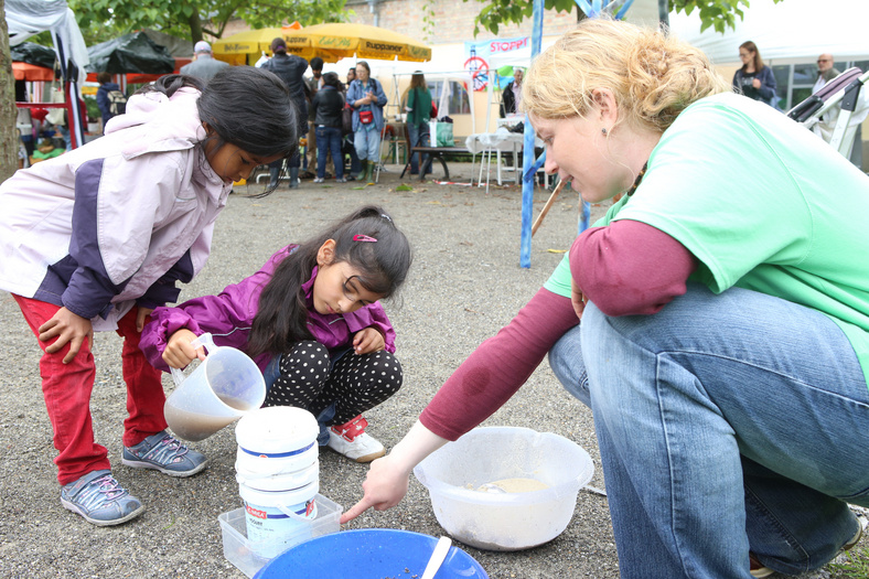 Nachhaltigkeitstage 2014 Zwei Kinder füllen eine Flüssigkeit in einen kleinen Eimer. Eine Schüssel steht daneben. Eine Erwachsene in der Hocke zeigt mit dem Finger auf den Füllstand des Eimers.