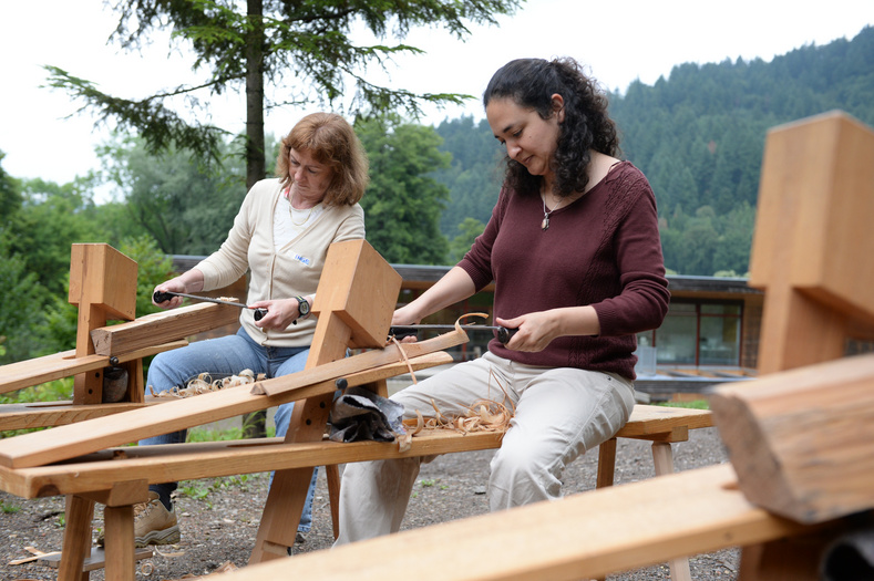 Zwei Frauen sitzen im Freien an Tischlerwerkzeugen und hobeln mit großen Messern eingeklämmte Hölzer.