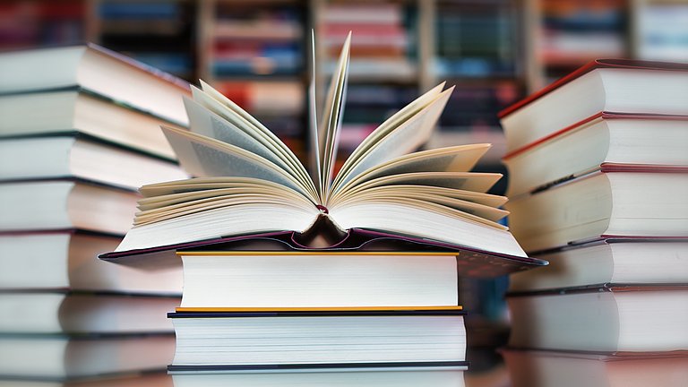 Im Zentrum des Bildes sieht man mehrere Bücher aufeinander. Im Hintergrund sind unscharf ebenso Bücher aufeinander gestapelt.