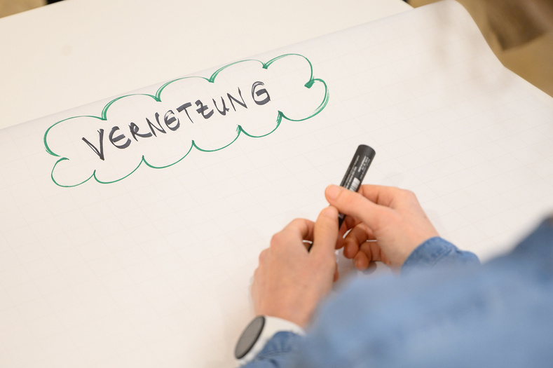 Das Bild zeigt einen Ausschnitt eines beschrifteten weißen Papiers, auf dem Vernetzung geschrieben steht und mit einem grünen Wellenkreis umrandet ist. Davor sieht man zwei Hände, die einen Stift in der Hand halten.