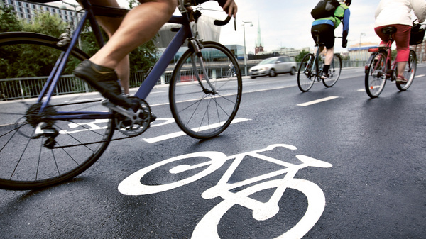 Dargestellt ist ein Fahrradweg parallel zu einer Straße, auf dem Radfahrer unterwegs sind.