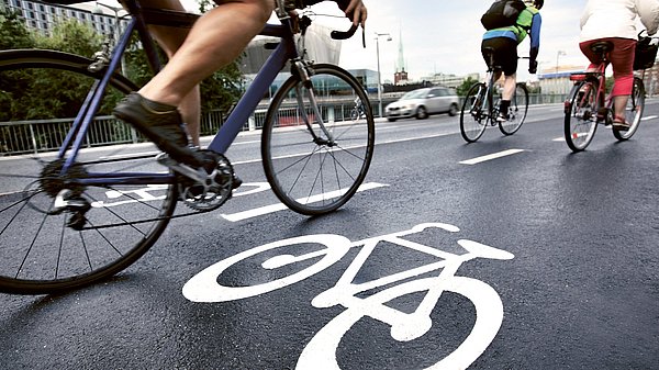 Dargestellt ist ein Fahrradweg parallel zu einer Straße, auf dem Radfahrer unterwegs sind.