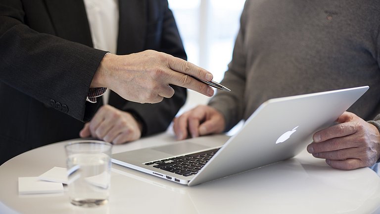 Im Mittelpunkt des Bildes befindet sich ein Laptop, weiter sind von zwei Personen der Rumpf zu sehen. Beide befassen sich mit dem Inhalt auf dem Bildschirm.