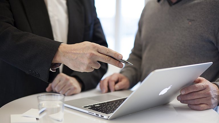 Im Mittelpunkt des Bildes befindet sich ein Laptop, weiter sind von zwei Personen der Rumpf zu sehen. Beide befassen sich mit dem Inhalt auf dem Bildschirm.