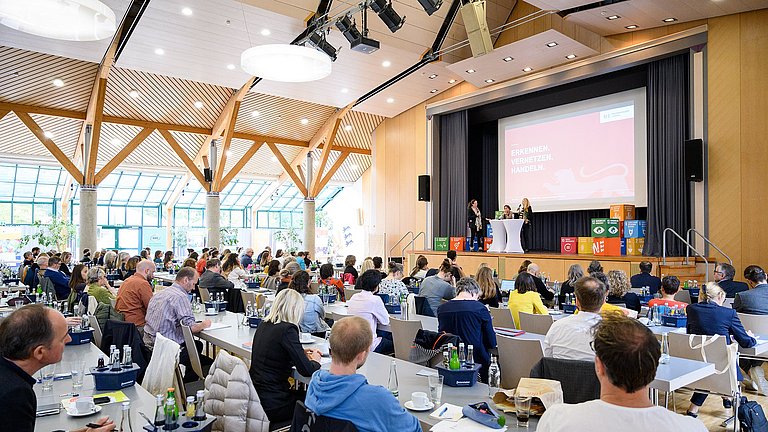 Das Bild zeigt eine Veranstaltungshalle, in der einige Personen an Tischen sitzen und den Blick auf die Bühne richten, die rechts hinten im Bild zu sehen ist. Dort stehen drei Frauen, darunter Kultusministerin Theresa Schopper und Umweltministerin Thekla Walker.