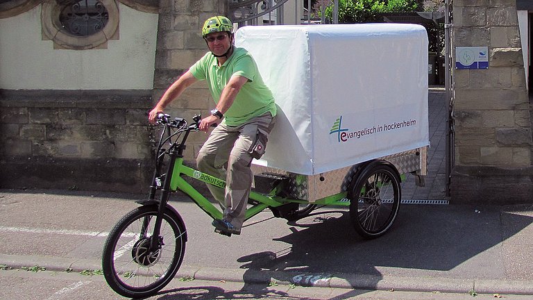 Ein Mann fährt mit einem Lastenfahrad, das hinten seine Ladefläche hat, aus einer Hofeinfahrt heraus.