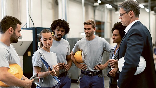 Dargestellt ist eine Szene aus dem Arbeitsleben. Eine Frau hält ein Klemmbrett in der Hand und zeigt Mitarbeiterinnen und Mitarbeitern etwas.